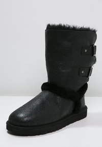 Botte en simili cuir noir avec une finition texturée, dotée d'un bord en fourrure noire douce et de deux boucles argentées pour la fermeture. Semelle plate noire.