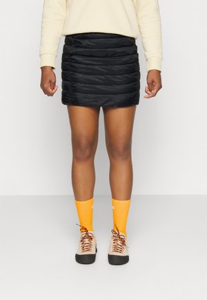 Persoon met een korte zwarte doorgestikte rok, crèmekleurig sweatshirt, feloranje sokken en beige wandelschoenen met zwarte zolen en meerkleurige veters.