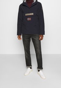 Męski model w granatowej kurtce z kapturem z naszywką przedstawiającą flagę Norwegii i logo, ciemnych dżinsach z mankietami oraz białych sneakersach, stojący na jednolitym tle.