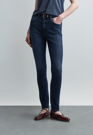 Persona che indossa jeans skinny blu scuro con cintura nera, maglietta grigio scuro e scarpe basse color bordeaux, in piedi davanti a uno sfondo grigio uniforme.