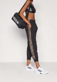 Černé sportovní kalhoty s leopardím potiskem na bocích, doplněné černou sportovní podprsenkou a bílými teniskami, nesoucí černou sportovní tašku adidas.