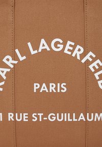 Rudas audinio maišelis su didelėmis baltomis raidėmis, rašančiomis "KARL LAGERFELD PARIS 1 RUE ST-GUILLAUME". Raidės viršuje yra išlenktos, o apačioje tiesios.