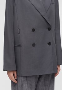 Blazer croisé gris foncé à quatre boutons noirs, une main reposant à côté d’un pantalon à carreaux noir et blanc.