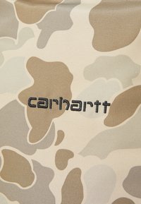 Tissu camouflage dans des tons beiges et marron clair avec des formes abstraites. Le logo "carhartt" est brodé en fil foncé.