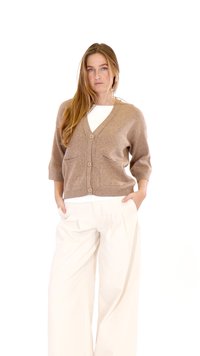 Kobieta stoi z rękami w kieszeniach, ubrana w beżowy rozpinany sweter na guziki, białe szerokie spodnie i biały top.