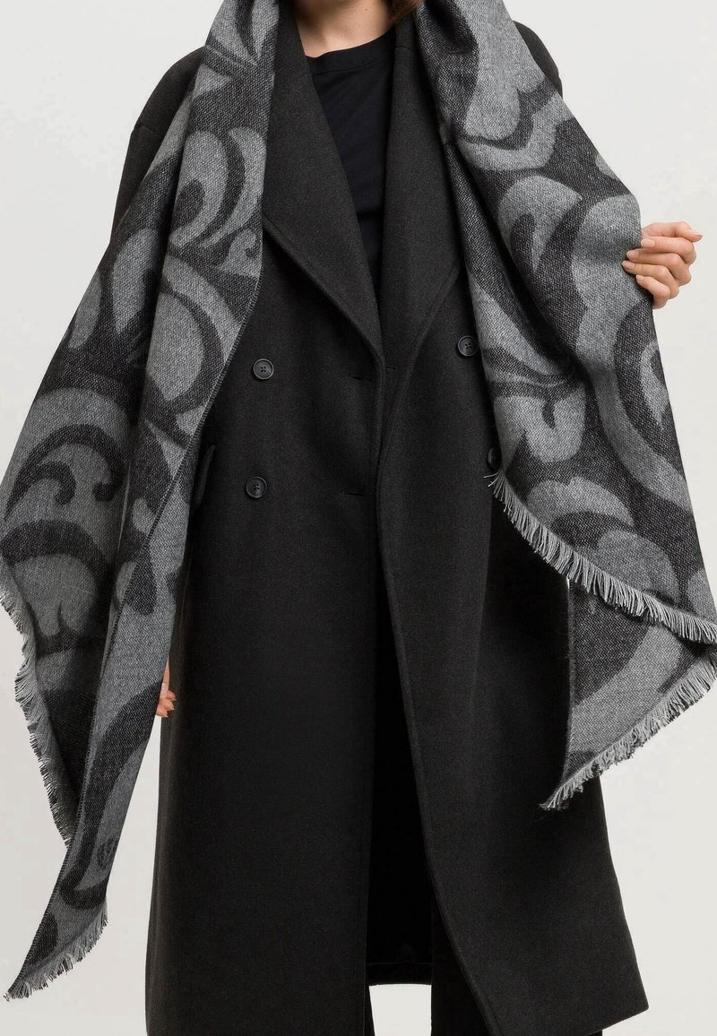 Personne portant un long manteau noir à double boutonnage tenant ouverte une grande écharpe grise avec des motifs abstraits foncés et des bords à franges.