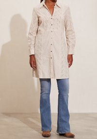Abito button-up beige chiaro a maniche lunghe con motivo floreale testurizzato e un leggero design ad occhio; abbinato a jeans a zampa d'elefante blu e stivali marroni.