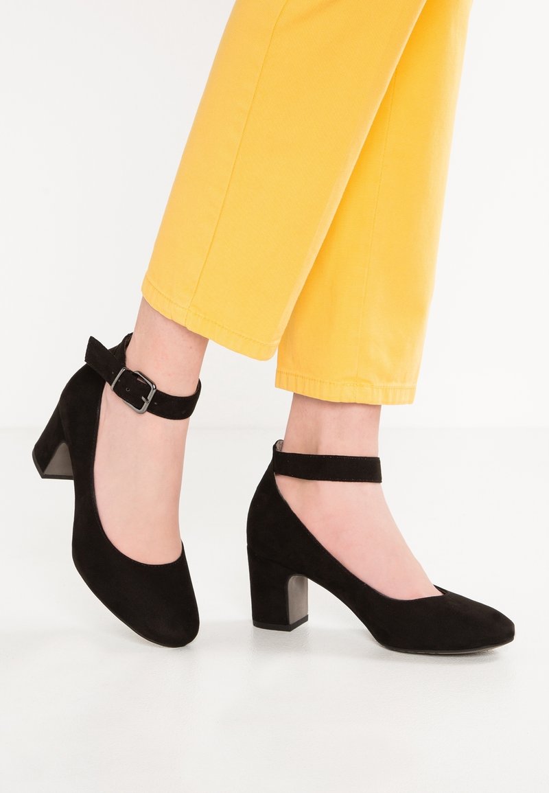 Pieds portant des chaussures Mary Jane en daim noir à talons blocs avec brides à la cheville, associés à un pantalon jaune raccourci sur fond blanc.