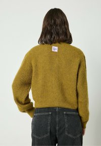 Gorčično rumena pletena jopica z rebrasto teksturo, z majhno etiketico na hrbtni strani. V kombinaciji s temno sivo hlačami.