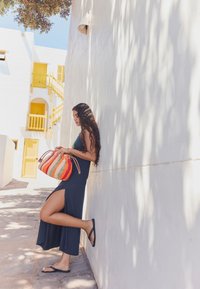 Robe longue bleu marine avec une fente haute sur la cuisse, associée à des tongs noires et un sac tissé multicolore. Mur blanc avec des accents jaunes en arrière-plan.