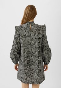 Nainen, jolla on pitkät hiukset, pukeutuneena mustaan mekkoon, jossa on valkoinen kukkakuosi, röyhelöillä varustetut olkapäät ja puhvihihoja, selin valkoista taustaa vasten.