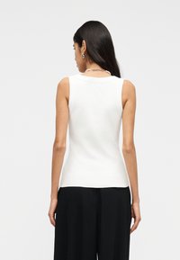 Mujer de cabello largo y oscuro, con pendientes plateados, un collar de perlas, una blusa blanca sin mangas y pantalones negros de pierna ancha, vista de espaldas.