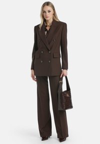 Blazer marrón de doble botonadura con bolsillos laterales, combinado con pantalones de pierna ancha a juego. Los accesorios incluyen un bolso oscuro con relieve.