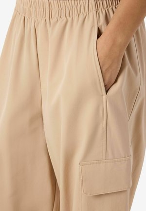 Pantaloni cargo bej cu talie elastică, buzunare laterale și un buzunar mare cu clapetă pe coapsă, confecționați dintr-un material neted.