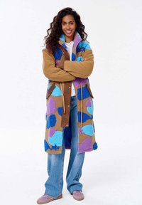 Kobieta z długimi kręconymi włosami stoi z założonymi rękami, ubrana w długi brązowy płaszcz z niebieskimi i fioletowymi puszystymi łatami, niebieskie dżinsy i różowe buty.