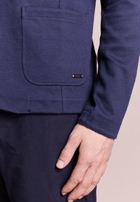 Námořnické modré texturované sako s přední kapsou a nenápadnou značkovou cedulkou. Zobrazuje ruku odpočívající na boku saka a ukazující detail manžety.