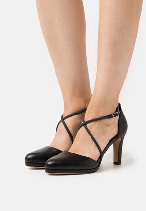 Escarpins noirs en cuir avec un bout pointu, une bride de cheville avec des détails croisés, et un talon fin. Texture lisse avec une finition mate.