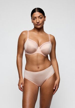 Vrouw met donker haar, gekleed in een bijpassende licht perzikkleurige beha en hoge taille slip, staand tegen een effen lichtgrijze achtergrond.