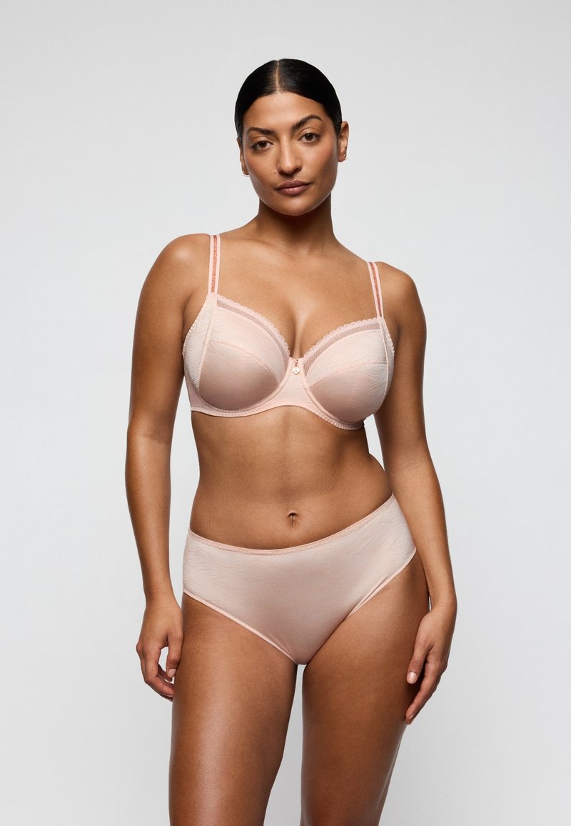 Tummahiuksinen nainen, joka pukeutuu vaalean persikan väriseen rintaliivipariin ja korkeavyötäröisiin alushousuihin, seisoo yksinkertaisen vaaleanharmaan taustan edessä.