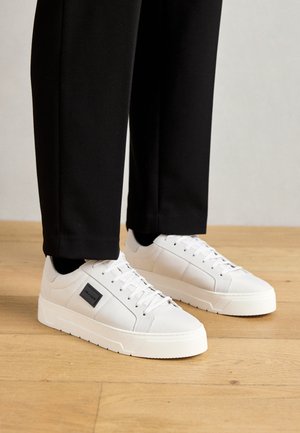 Weiße Sneakers mit einem glatten Obermaterial aus Canvas, einem schwarzen Logo-Patch und einer dicken weißen Gummisohle. Kombiniert mit schwarzen Hosen.