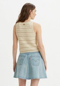 Donna che indossa un top senza maniche in crochet beige e una gonna di jeans azzurri, vista di schiena su uno sfondo bianco uniforme.