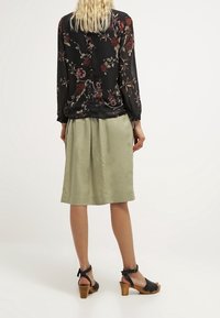 Blusa nera floreale con maniche lunghe, abbinata a una gonna plissettata verde chiaro e sandali neri con tacco, vista da dietro.