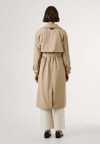 Person klädd i en beige trenchcoat med bälte, vida vita byxor och svarta skor, som står med ryggen mot en enkel bakgrund.