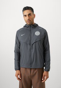 Chaqueta Nike gris con cierre frontal, capucha y logotipo en el pecho. Presenta un diseño estampado, mangas largas y corte recto.