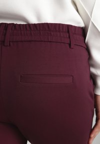 Primer plano de una persona que lleva pantalones burdeos con un bolsillo posterior de ojal y una cinturilla elástica, combinados con una blusa blanca.