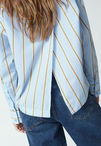Himmelblau gestreiftes Hemd mit gelben Streifen, einem Schlitz im Rücken und langen Ärmeln. Kombiniert mit dunklen Jeans.