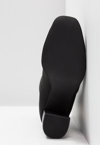 Semelle d'une botte noire à talon carré reposant contre un fond d'angle blanc, montrant une prise texturée à la base du talon.