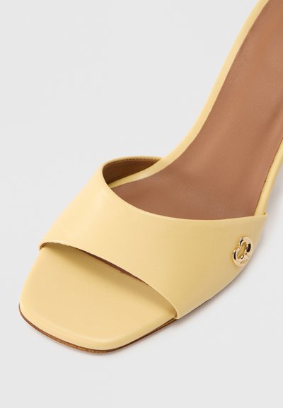 Κίτρινο πέδιλο με τακούνι, κατασκευασμένο από λείο δέρμα με ανοιχτή μύτη. Διαθέτει ευρύ, καμπυλωτό λουράκι μπροστά και δια κοσμητικό λογότυπο σε χρυσό τόνο.