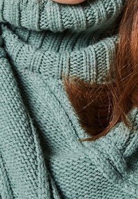 Teksturowany, dzianinowy sweter w miękkim zielonym kolorze z wysokim, prążkowanym golfem. Gładki materiał luźno opada, ukazując skomplikowane wzory i fałdy.