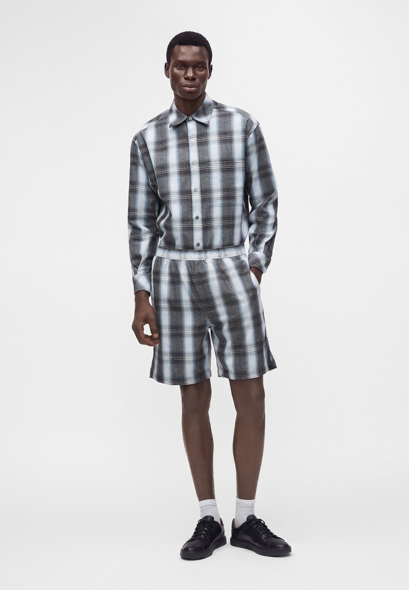 Homme debout les mains dans les poches portant une chemise à carreaux bleue et noire boutonnée et un short assorti à carreaux avec des chaussures noires et des chaussettes blanches.