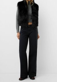 Mustat tekoturkishaluttimen harmaan poolopaitapuseron päällä, yhdistettynä leveälahkeisiin mustiin farkkuihin, joissa on hopeisia niittej ä sivuilla. Ruskeat nilkkurit viimeistelevät asun.