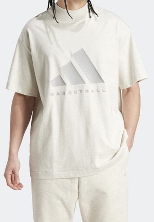 Persona che indossa una T-shirt oversize beige chiaro Adidas e pantaloncini abbinati con il logo Adidas Basketball sul petto.