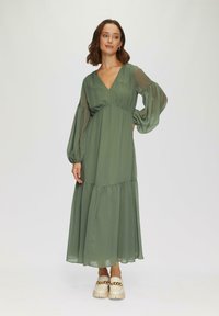 Robe longue en chiffon vert avec décolleté en V, jupe superposée, manches bouffantes et design fluide, accompagnée de chaussures à plateforme chunky crème.