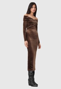 Brązowa aksamitna sukienka z dekoltem odkrywającym ramiona, długimi rękawami i marszczonymi detalami; dopasowany krój sięgający do połowy łydki. Czarne kozaki za kolano.