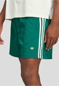 Grønne atletiske shorts i et let materiale, med hvide tre striber ned langs siderne og et lille guld-logo på kanten.