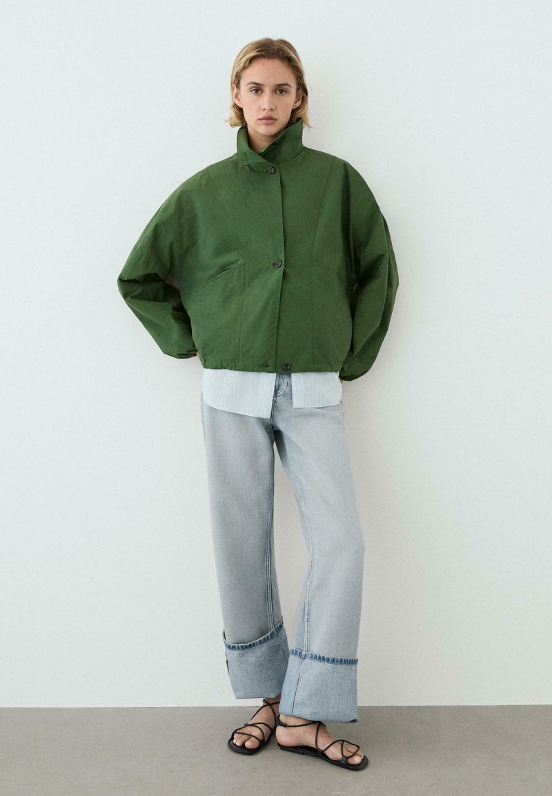 Vrouw staat tegen een witte muur, draagt een groen jasje met knopen, lichtblauwe wijde omslagspijpen jeans en zwarte sandalen met bandjes.