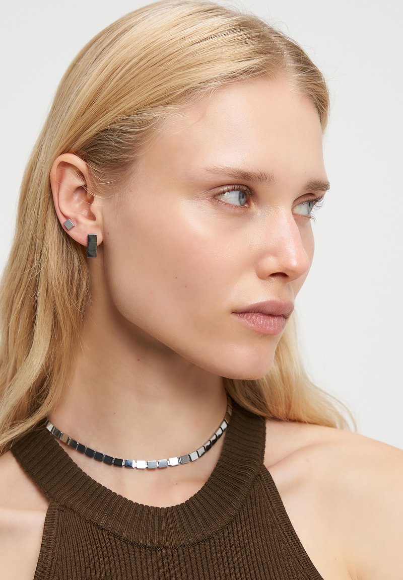 Blonde Frau in einem dunklen ärmellosen Oberteil, mit quadratischen silbernen Ohrringen und passendem quadratischen metallischen Choker, die nach rechts auf einem weißen Hintergrund schaut.