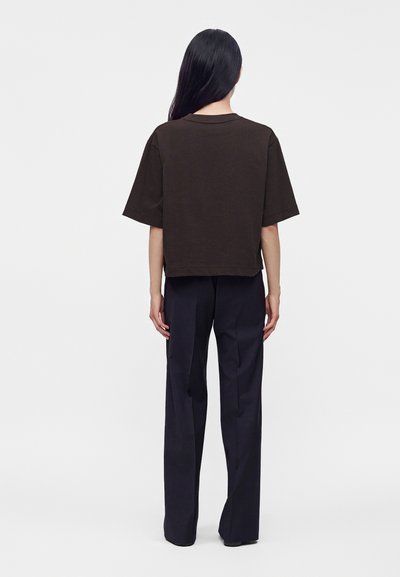 Personne aux longs cheveux noirs, de dos, vêtue d'une chemise noire ample à manches courtes et d'un pantalon noir droit sur un fond uni.
