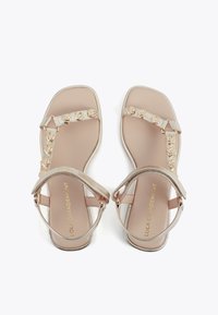 Sandales plates beiges avec brides à la cheville et clous décoratifs dorés, vues de dessus sur fond blanc.