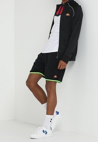 Person i sort sportsjakke og shorts med neongrønne detaljer, hvid polo t-shirt, hvide sokker og hvide sneakers med blå accenter.