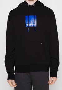 Sudadera negra de tela suave, con un logo cuadrado azul en el pecho, capucha con cordones y un bolsillo tipo canguro en la parte frontal.
