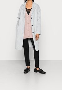 Žena v světle šedém kabátu, růžovém svetr s knoflíky, černém topu s krajkovým lemem, černých slim pants a černých mokasínech s mašlemi.