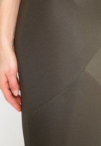 Robe ajustée texturée en vert olive foncé avec des détails de coutures diagonales et un motif en tissu côtelé ; main reposant sur le côté pour l'échelle.