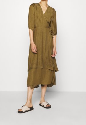 Frau trägt ein olivgrünes Wickelkleid mit ellenlangen Ärmeln und gestuftem Saum, kombiniert mit beigen Pantoletten vor einfarbigem Hintergrund.