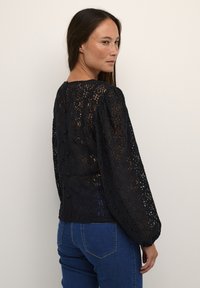 Sort blonde med lange pufærmer, blomster mønster, rund halsudskæring og knapdetaljer på ryggen, båret med blå jeans.