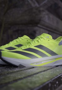 Par neon gule Adidas løbesko med sorte striber og hvide såler, hvilende på en træoverflade udendørs.